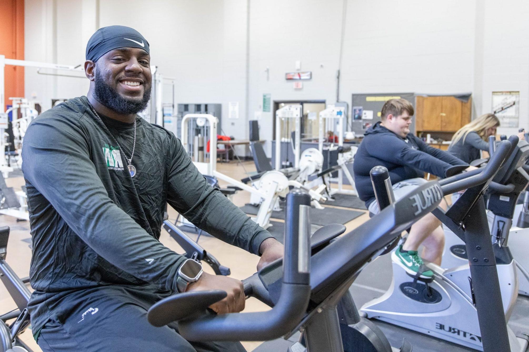 A man on a cycling machine in a fitness center with two other people also on cycling machines behind him.